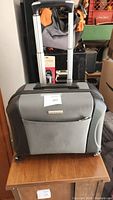 Grey Samsonite carry on suitcase with front pocket and extended telescopic handle, placed on wooden table with background of other items.