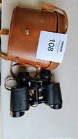 Moon Beam binoculars with textured grips next to brown leather case with shoulder strap.