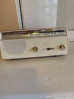 Front angle view showing the cream-colored rectangular radio with perforated speaker grill, gold-tone trim, two gold rotary knobs, and tuning scale.
