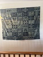 Full view of the Nigerian batik wall fabric hanging showing entire geometric pattern composed of squares and rectangles with different motifs in varied shades of blue, gray, and black.