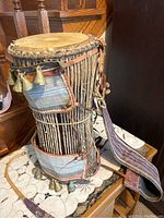 Full view of the Nigerian talking drum showing the wooden body, fabric sash, and attached brass bells.