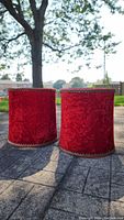 Pair of red crushed velvet barrel lampshades with gold trim standing outside on pavement, showing front sides.