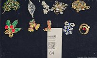 Top-down view of 11 vintage holiday and rhinestone brooch pins on black fabric background showing various shapes and colors including holly, bells, snowflakes, floral and abstract designs.