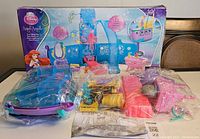 Photo showing the full box with Disney Princess Ariel themed 2 in 1 Royal Ship playset pieces sealed in plastic bags, including blue sails, pink vanity, and other parts on a table.