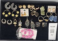 Overhead photo showing a black tray with mixed vintage costume jewelry earrings including plastic hoops, gold-tone ornamental earrings, fabric clip-ons, and beaded pairs.