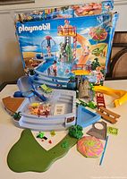 Full overview of Playmobil beach set pieces arranged showing main pool, slides, umbrella, base pieces, and box in background.