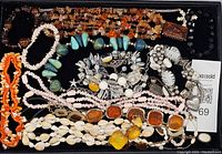 Top-down image showing a collection of vintage costume jewelry including bead necklaces, shell necklaces, silver-tone brooches with rhinestones, and a few bracelets laid out on a black surface.