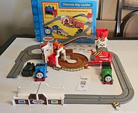 Overview of the Thomas & Friends Big Loader playset with most components arranged on a flat surface, including trains, tracks, loader, fence pieces, and box in background.