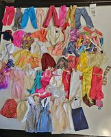 Full display of assorted vintage Barbie and fashion doll clothing laid out on a white surface showing pants, tops, dresses, skirts, jackets, and bag of accessories.