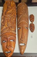 Two large carved wooden masks and three smaller tribal wooden masks displayed together on a flat surface. Larger masks approximately 40 and 36.5 inches tall, smaller masks around 6.5 inches.