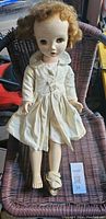 Full shot of vintage doll seated on wicker chair showing one foot bare, the other foot with a damaged shoe and sock. Doll wearing a white dress, head attached but showing dirt and wear.