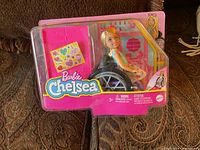 Front view of Barbie Chelsea doll seated in wheelchair inside original pink-branded packaging.