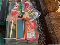 Photo of bin containing Barbie and Barbie-like dolls and some miniature furniture items such as a blue doll bed frame and plastic bags with accessories.