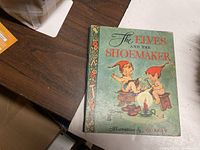 Front cover of the children's book 'The Elves and the Shoemaker' showing illustrated elves on green background with title and illustrator name Dorcas.