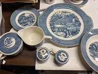 Photo showing cake plate with Rocky Mountains design, creamer, sugar bowl with lid, salt and pepper shakers, and some bread plates on dark wooden surface.