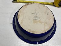 Bottom of blue ceramic pie plate showing Emile Henry stamp and approximate size measurement with tape measure.
