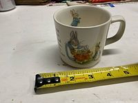 Side view of the child’s ceramic mug featuring an image of Peter Rabbit stirring a pot, next to a measuring tape showing approximate size.