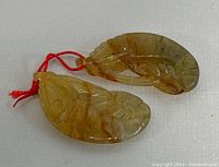 Two carved jade pendants shaped like fish/shrimp on red cords lying flat on white background.