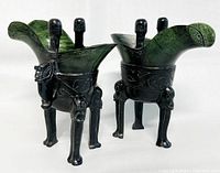 Pair of carved jade liquor cups with dragon designs on black metal stands, viewed at angle showing details of carving and stands.