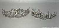 Two silver-tone wedding crowns displayed side by side on a plain surface, highlighting their rhinestone and pearl-like bead designs.