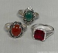 Front view of three silver rings showing colored gemstones: greenish-blue, orangish-red, and square red gem.