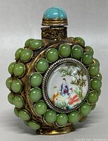 Front and side view of vintage Chinese snuff bottle showing turquoise cabochon stopper, green cabochon decoration on brass/bronze body, and one painted porcelain roundel with traditional Chinese figures and landscape.