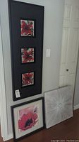 Vertical black-framed triptych painting featuring three red flowers on canvas beside two square art pieces on floor.