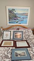Overview of six framed artworks and one empty frame on bed with floral patterned bedspread