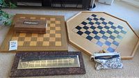 Wide shot showing a wooden chess board, octagonal checkers board, cribbage board with metallic peg section and fabric pouch with pegs, all arranged on floor.