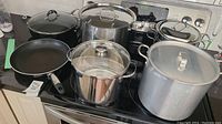 Overview of six pots and pans including non-stick frying pans and aluminum and stainless steel pots with glass lids on a stove.