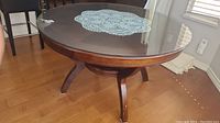 View of round kitchen table side showing curved solid wood legs with dark finish and glass top with crocheted doily underneath in natural light.