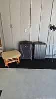 Photo showing two pieces of luggage, two walking sticks, and a cushioned ottoman against a wall with cupboards.