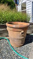 Large terracotta planter pot outdoors in a garden bed with nearby plants. Shows the aged weathered surface and ring and handle detail.