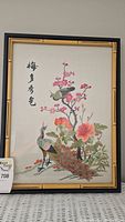 Framed artwork depicting two peacocks under a blooming tree branch with flowers and a smaller bird, with Chinese calligraphy, held upright for photo