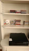 Photo of the Sony CD player on shelf above white cabinet, with multiple CDs and cassette tapes on shelves above