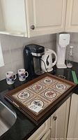 Photo showing black single-serve coffee maker, white electric kettle, white SodaStream machine with bottle, two floral mugs, and wooden tile tray on countertop near kitchen sink.