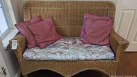Wicker loveseat with patterned seat cushion and four pink throw pillows placed on it, showing overall condition and structure.