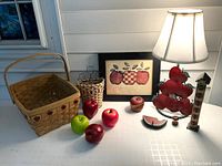 Overview of lot items including framed print, multiple apple motif baskets, assorted apple figurines, apple lamp base, and small wooden stool with apple-shaped seat in bright red tones.