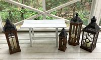 Five Moroccan style lanterns made from wood and perforated metal with glass panels, each containing a battery-powered candle, arranged on porch floor with white bench in background.