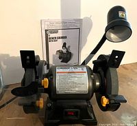 Front view of the 6-inch bench grinder with light and owner's manual, showing wheel guards, eye shields, power switch, and flexible goose neck light.