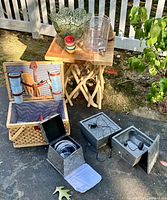Photo showing the picnic basket open with cloth lining, cups attached to lid, wooden salad tongs, colorful small bowls, large plastic salad bowl, lemonade dispenser, and two slate tabletop fountains with cords.