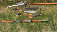 Overview of all yard tools laid out on grass showing seven items: Fiskars hatchet, anvil lopper, folding saw, hand pruners, machete with sheath, extendable tree pruner, and Ridgid bow rake.