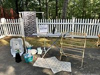 Full lot viewed outdoors against white picket fence, showing folding ironing board with cover, gold-tone drying rack, laundry basket, foot mats, Sunbeam iron, and various laundry and home care items.