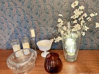 Wide view of the various glass items in the lot including the clear jellyfish light inside a tall clear vase, footed milk glass bowl, clear glass globe, electric candles and amber glass vase placed on table