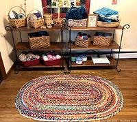 Photo showing two wood and wrought-iron shelving units loaded with baskets of yarn, storage boxes, folded fabrics, and a colorful braided rag rug in front.