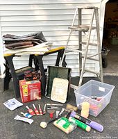 Lot of painter's supplies including sawhorses, step ladder, various tarps, paint trays, brushes, rollers, and tools on an outdoor surface