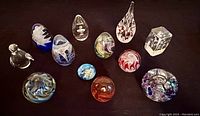 Overall view of all 12 glass paperweights arranged on a dark surface showing various shapes and colors including spheres, teardrops, bird figure, and cube.