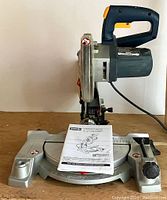 Side view of Ryobi corded mitre saw with manual on base showing handle, base, and blade guard.