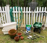 Garden items including seed spreader, pots, stakes, cages, hose, hand tools, rake, shovel, potting mix and watering can grouped on grass in front of white picket fence