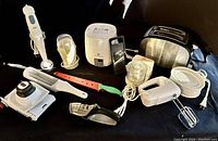 Photo showing all nine items in the lot on a dark background including the Braun whisk, rice cooker, grinder, hand mixer, chopper, sharpener, bagel separator, watermelon cutter, and Zyliss tool.
